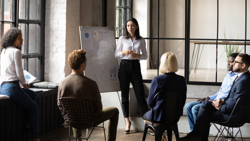 Standing speaker briefs a small group using a marker and flip chart in an industrial-style room.