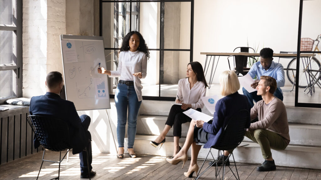 Presenter explains diagrams on a flip chart to seated colleagues in a sunlit workspace.