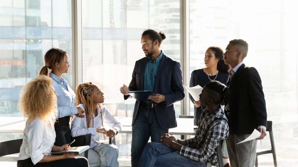 Team gathers around a leader with a clipboard for an informal discussion in a bright loft.
