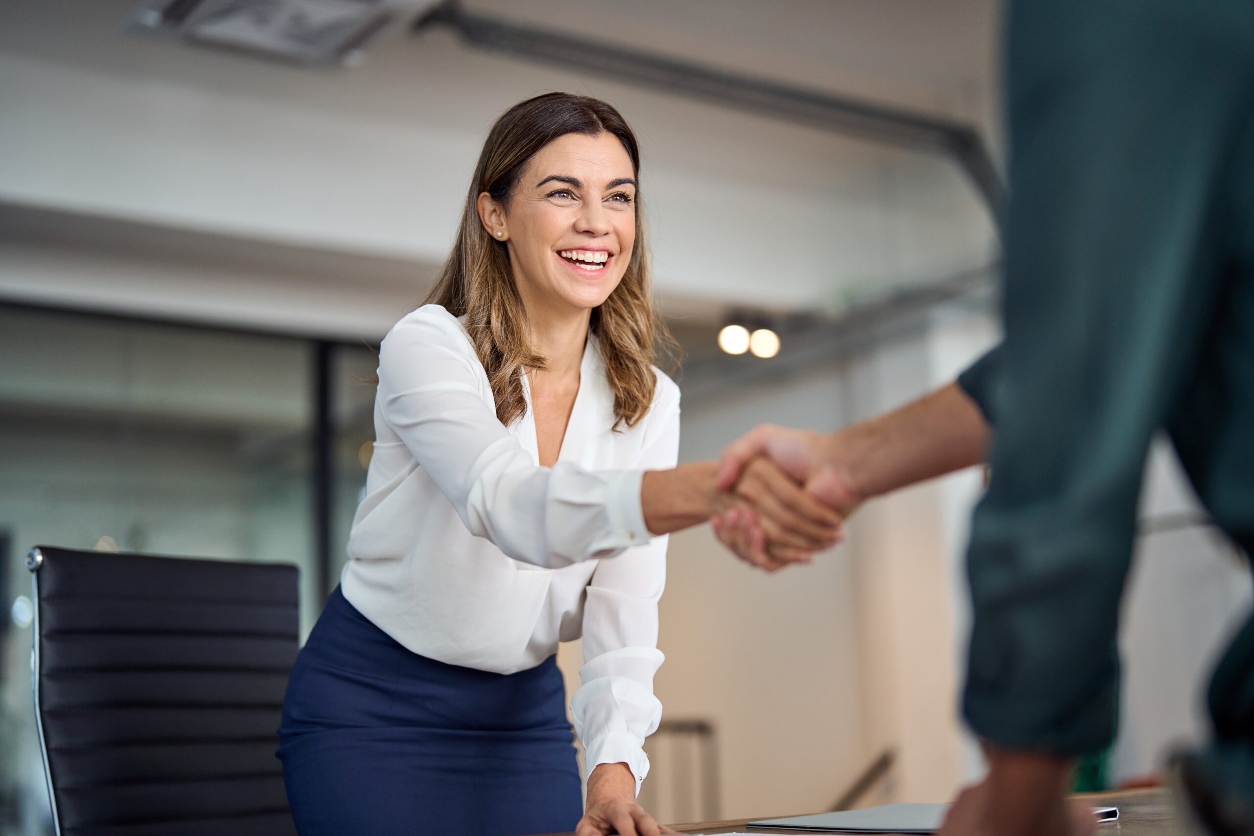 Smiling executive leans forward to seal a deal with a handshake in a modern office.
