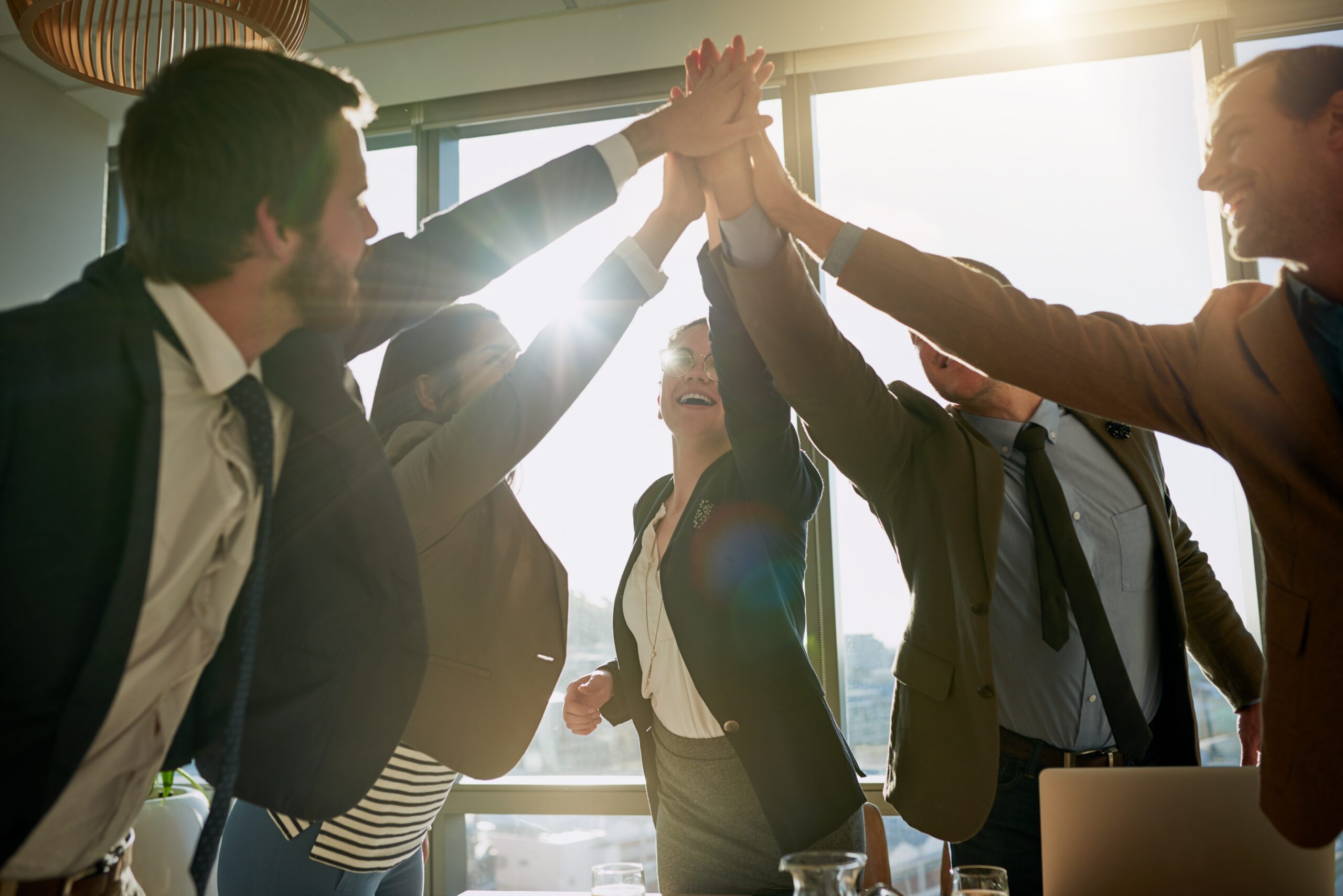 Business team celebrates a win with a group high-five backlit by sunlight.