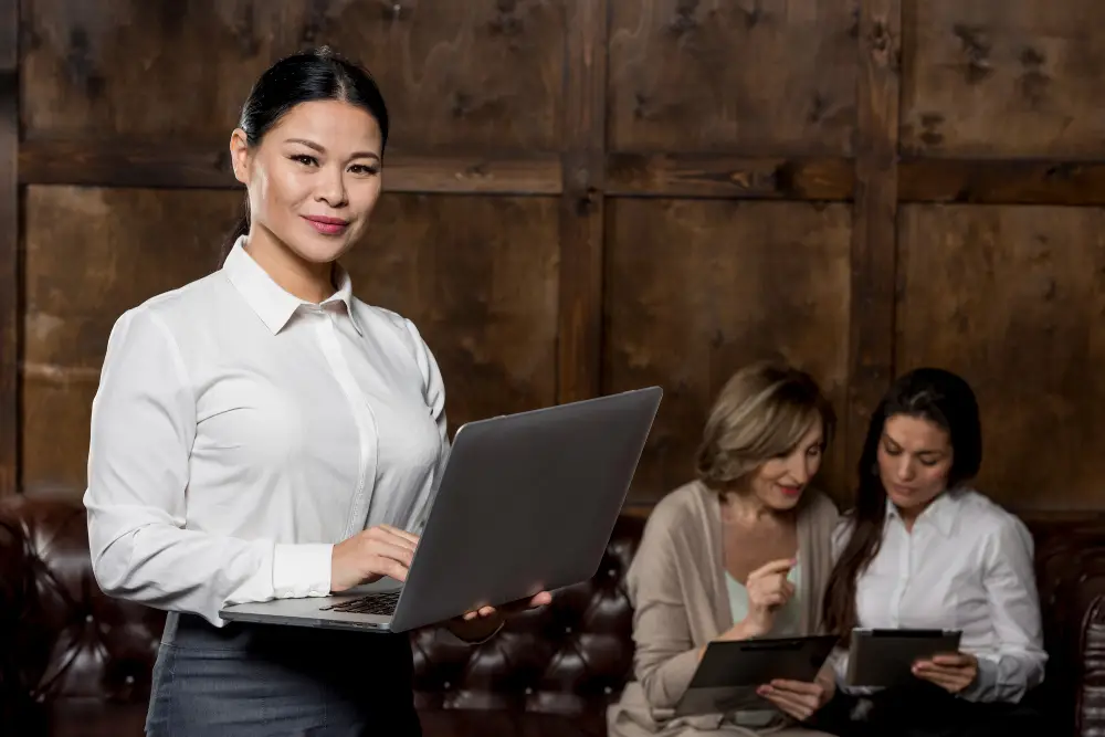 Confident professional stands with a laptop while two colleagues review tablets behind her.