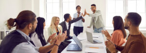 Mixed race business team celebrating good results in office meeting. Happy young black man gives high five to coworker while diverse multi ethnic teammates are applauding. Teamwork and success concept
