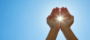Hands cupped toward the sky capture bright sunlight streaming between the palms.