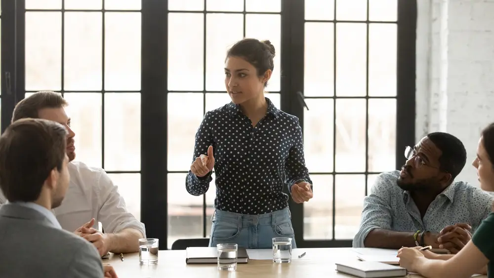 Young woman leads a meeting, gesturing as teammates listen at a sunlit conference table.