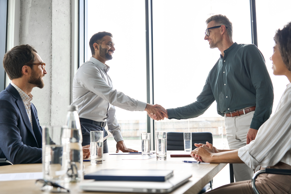 Two businessmen lean across a conference table to shake hands as others look on approvingly.