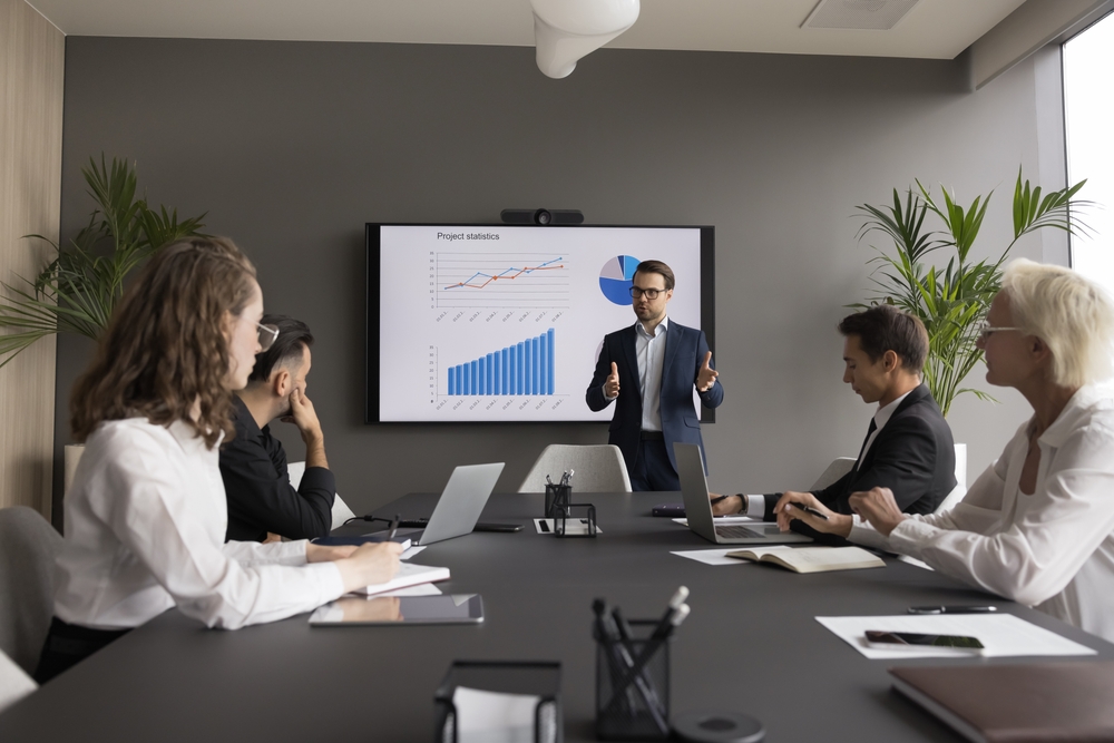 Executive presents project statistics on a screen to colleagues gathered around a boardroom table.