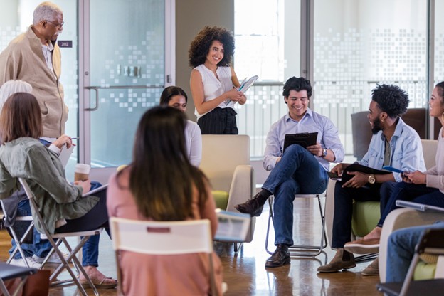 Facilitator stands with a tablet while participants in a circle share ideas in a modern meeting space.