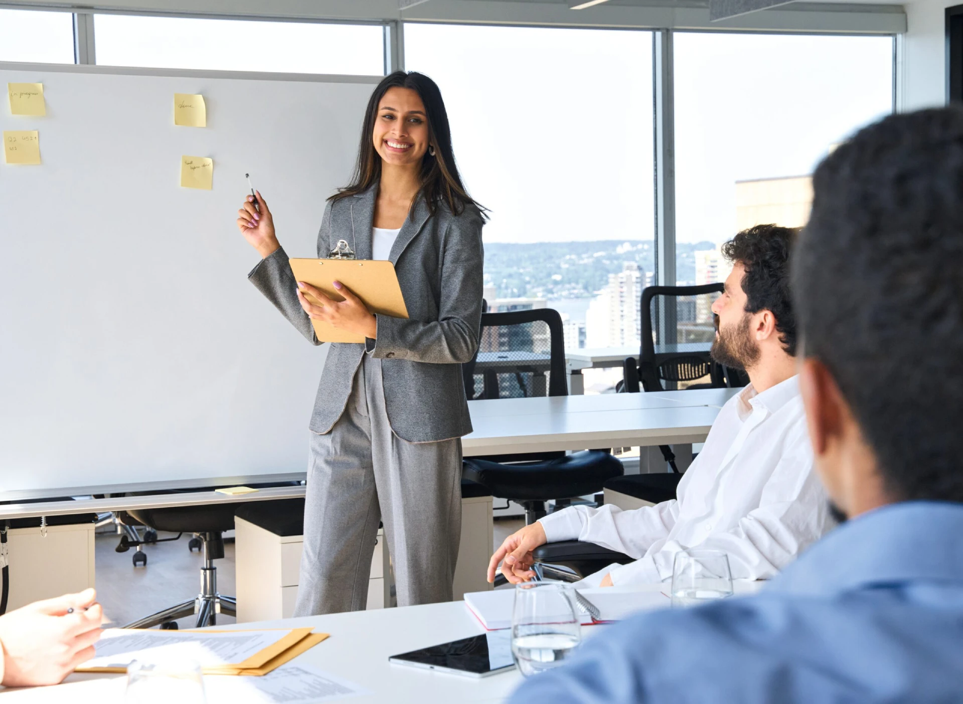 Presenter with clipboard leads a strategy session, pointing at sticky notes on a whiteboard as the team looks on.